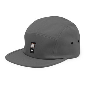 May include: A gray five-panel cap with a flat brim. The cap features a small embroidered design on the front, with a black square and a design inside. The cap has two black eyelets on each side.