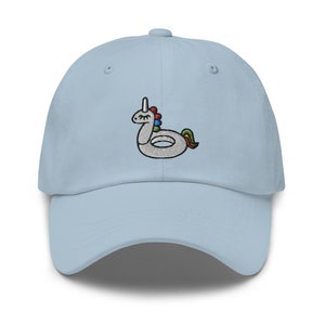 Può includere: Un cappello da baseball blu chiaro con un design ricamato di un unicorno gonfiabile. L'unicorno è bianco con una criniera e una coda arcobaleno.