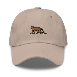 Può includere: Un cappello da baseball beige con un design di scimmia marrone ricamato.