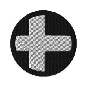 Puede incluir: Un signo más blanco sobre un fondo circular negro. El signo más está bordado con una tela texturizada.