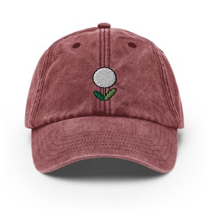 May include: A maroon vintage-style baseball cap with an embroidered white dandelion design.