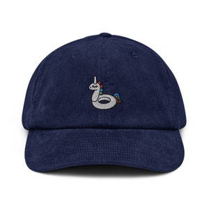 Può includere: Un cappello da baseball in velluto a coste blu navy con un design ricamato di un unicorno gonfiabile bianco.