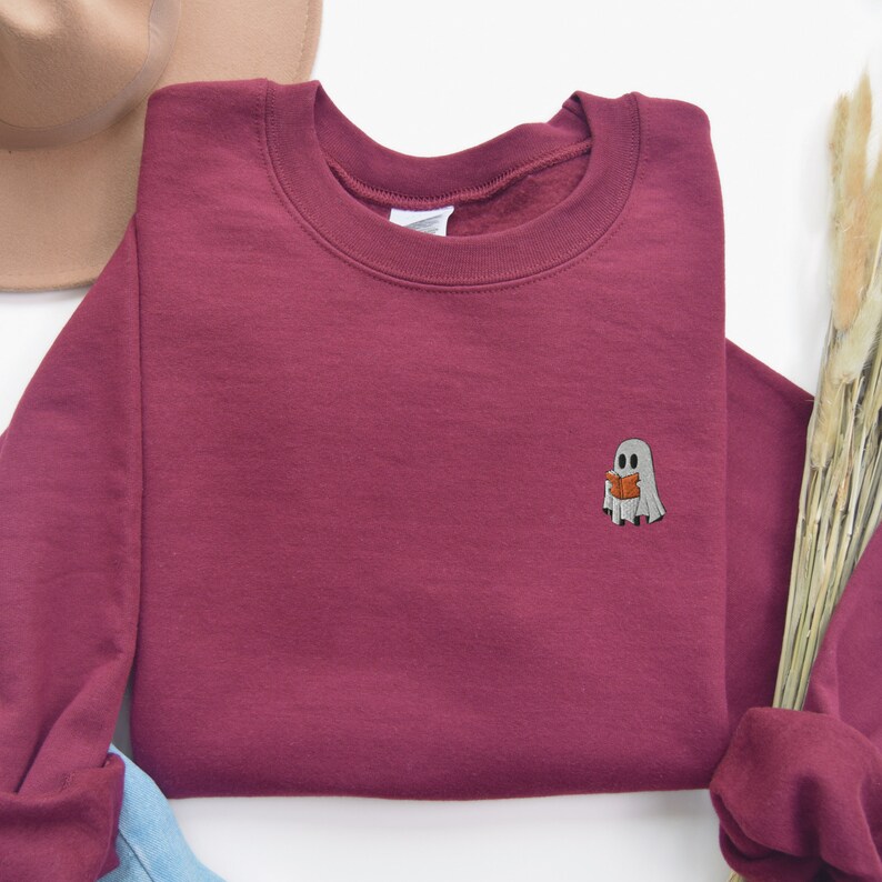 May include: A maroon sweatshirt with a small embroidered ghost holding a book. The ghost is white with black eyes and a black mouth.