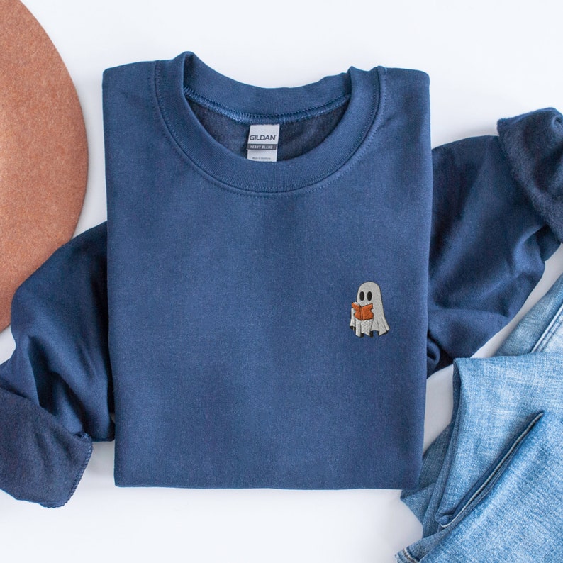 May include: A navy blue sweatshirt with a small white ghost reading a book embroidered on the front. The sweatshirt is folded and laid flat on a white surface.