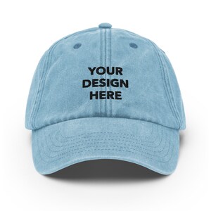 May include: A light blue denim baseball cap with the text "YOUR DESIGN HERE" embroidered in black on the front.