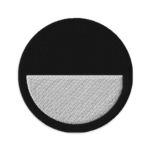 May include: A circular black embroidered patch with a white, semi-circular design at the bottom. The patch has a textured appearance, with the black portion appearing solid and the white section showing a diagonal pattern.