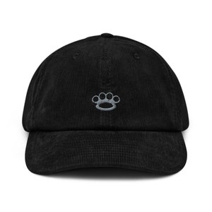 May include: Black corduroy baseball cap featuring a silver embroidered knuckle duster emblem. The cap has a curved peak and an adjustable strap. A fashionable accessory for everyday wear.