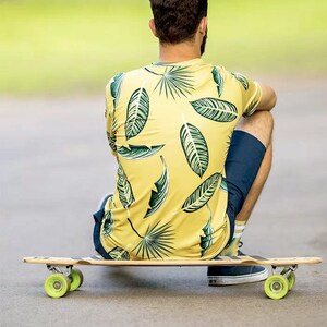 Puede incluir: Una persona sentada en una tabla de skate larga con una camisa de estampado tropical de hojas verdes y amarillas y pantalones cortos azul oscuro.