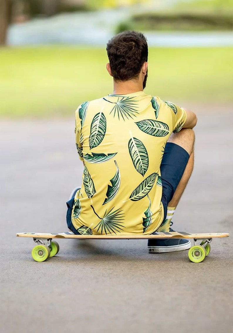 Puede incluir: Una persona sentada en una tabla de skate larga con una camisa de estampado tropical de hojas verdes y amarillas y pantalones cortos azul oscuro.