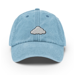 Puede incluir: Una gorra de béisbol de denim azul claro con un diseño de nube bordada en blanco.