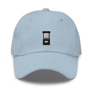 May include: A light blue baseball cap with an embroidered image of a vintage arcade game. The game has a black cabinet with a brown screen and a black joystick.