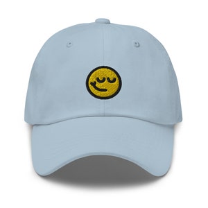 May include: A light blue baseball cap with a yellow smiley face embroidered on the front. The smiley face has closed eyes and a slight smile.