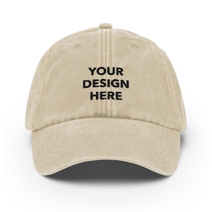May include: A beige cotton baseball cap with the text "YOUR DESIGN HERE" embroidered in black on the front.