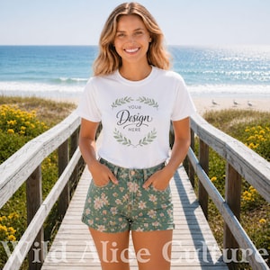 May include: A white t-shirt with the text "Your Design Here" inside a green wreath. The model is wearing green floral shorts. The background shows a wooden walkway, the ocean, and the sky.