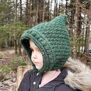 Può includere: Un cappello da folletto verde all'uncinetto con la parte superiore a punta e paraorecchie. Il cappello è realizzato con un filato strutturato e viene indossato da un bambino. Lo sfondo presenta una scena di foresta innevata.