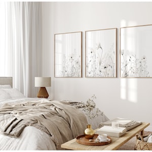 May include: Three framed black and white botanical prints of wildflowers with a light brown wood frame. The prints are hanging on a white wall above a bed with a beige blanket and a wooden coffee table.