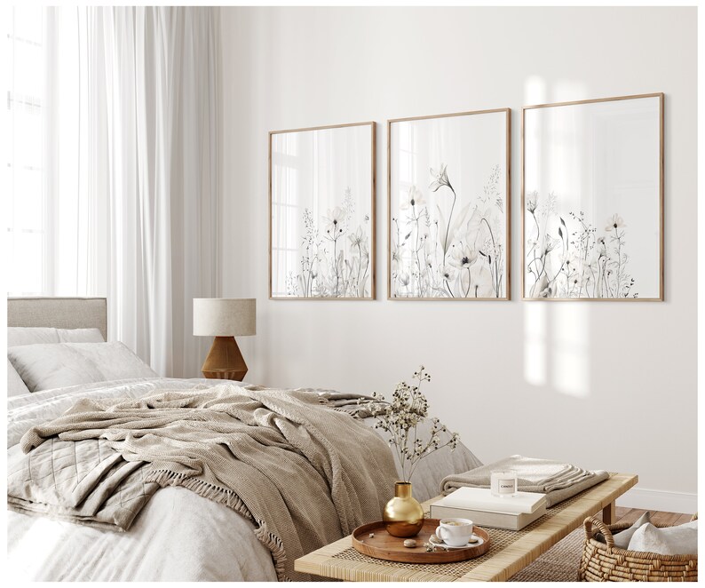May include: Three framed black and white botanical prints of wildflowers with a light brown wood frame. The prints are hanging on a white wall above a bed with a beige blanket and a wooden coffee table.