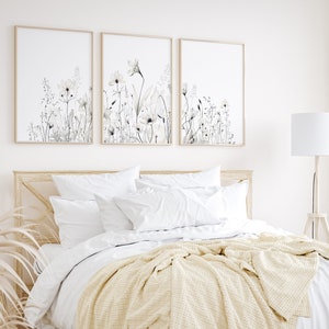 May include: Three framed prints of watercolor floral illustrations in shades of gray and white, hanging above a bed with white bedding and a cream throw blanket.