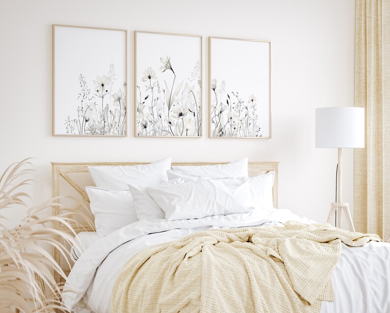 May include: Three framed prints of watercolor floral illustrations in shades of gray and white, hanging above a bed with white bedding and a cream throw blanket.