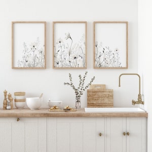 May include: Three framed prints of watercolor floral illustrations in shades of gray and white, hanging on a white wall above a kitchen counter with a wooden countertop and white cabinets. A vase with a sprig of greenery sits on the counter.