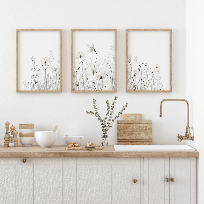 May include: Three framed prints of watercolor floral illustrations in shades of gray and white, hanging on a white wall above a kitchen counter with a wooden countertop and white cabinets. A vase with a sprig of greenery sits on the counter.
