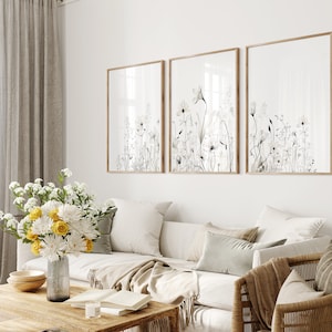 May include: Three framed prints of black and white watercolor floral illustrations with a white background. The prints are hanging on a white wall above a white couch with beige throw pillows and a brown blanket.