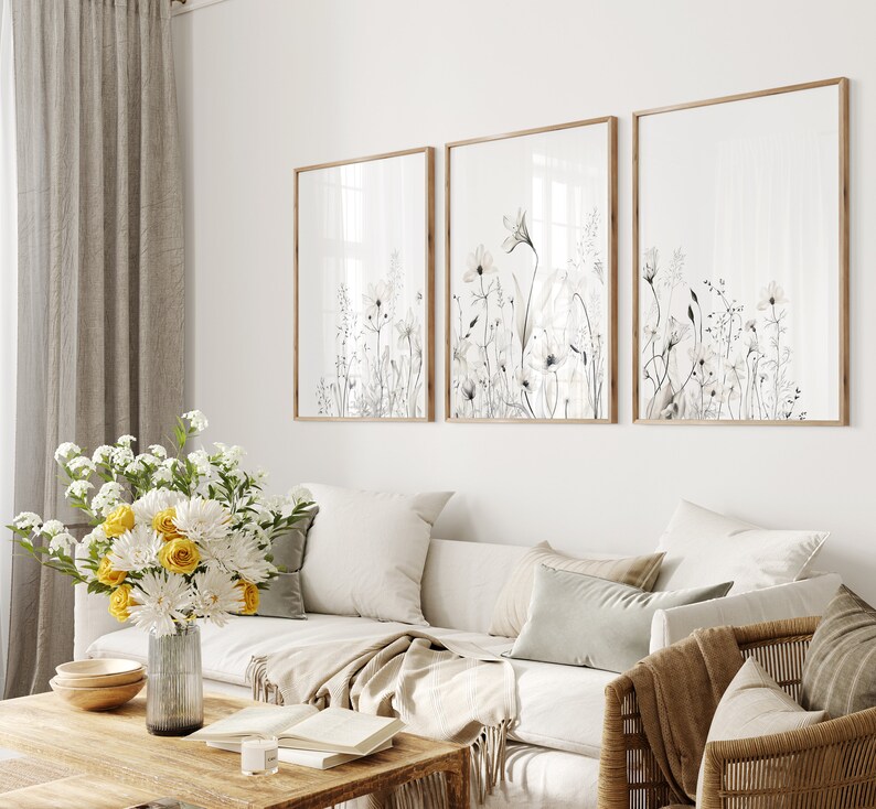 May include: Three framed prints of black and white watercolor floral illustrations with a white background. The prints are hanging on a white wall above a white couch with beige throw pillows and a brown blanket.