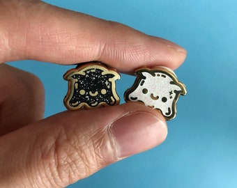 Puede incluir: Dos pines de esmalte de color dorado con forma de fantasmas. Un fantasma es negro con purpurina y el otro es blanco con purpurina.