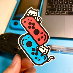 May include: Two blue and red Nintendo Switch Joy-Con controllers with cute white cat stickers. The cats are holding the controllers and have stars on their bellies.