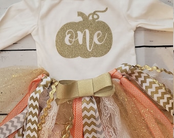 May include: A white long-sleeved onesie with a gold glitter pumpkin and the word "one" on the front. The onesie is paired with a tulle skirt with gold, peach, and gray chevron ribbons and a gold bow.
