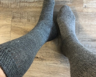 May include: Two gray knitted socks, one on each foot, are visible against a wooden floor background. The socks are a solid gray color and appear to be made of a thick, warm material. The socks are pulled up to the mid-calf.