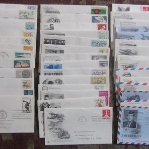 May include: A collection of vintage envelopes, many with postage stamps and postmarks. Some envelopes feature images of historical figures, airplanes, and other designs. The envelopes are stacked in three columns, showcasing a variety of postal ephemera.