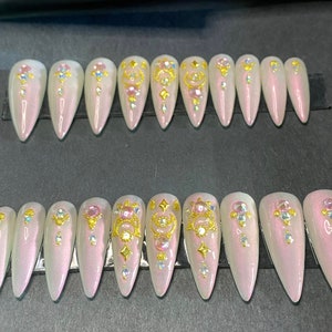 Puede incluir: Conjunto de 20 uñas postizas largas, blancas, iridiscentes, en forma de stiletto con adornos dorados y rosados.