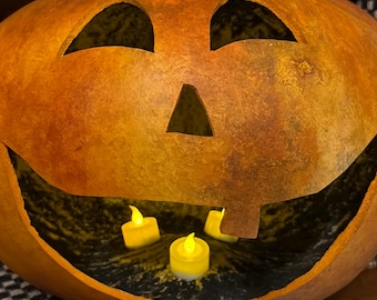Puede incluir: Una calabaza tallada con forma de linterna con un exterior texturizado y marrón moteado. El rostro presenta dos ojos triangulares y una boca ancha y abierta. Dentro del interior hueco, brillan tres pequeñas velas LED a pilas.
