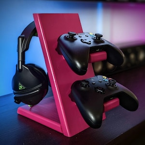 May include: A pink wooden stand with two black video game controllers and a black headset. The stand is designed to hold the controllers and headset upright.
