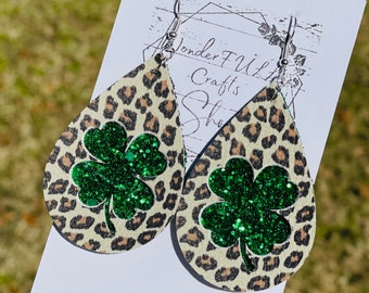 May include: A pair of dangle earrings with a leopard print background and a green glitter four-leaf clover design. The earrings are made of leather and have silver hooks.