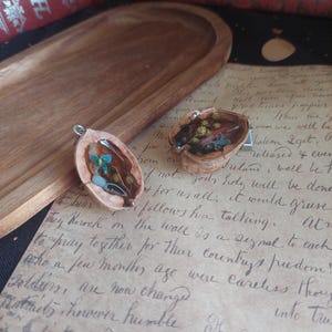 Peut inclure: Deux pendentifs en forme de coquille de noix remplis de résine et de fleurs séchées. L'un présente un papillon bleu et des feuilles vertes, l'autre des accents jaunes et rouges. Les pendentifs sont sur un fond de papier vintage.