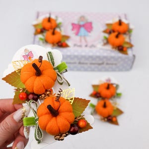 Könnte beinhalten: Nahaufnahme von dekorativem Haar-Accessoire zum Thema Herbst mit kleinen orangefarbenen Kürbissen mit braunen Stielen, grünen Blättern und Beeren. Die Accessoires sind auf weißem Karton mit einem dekorativen Design befestigt. Eine Schachtel mit ähnlichen Dekorationen befindet sich im Hintergrund.