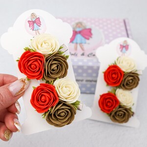 May include: Two hair clips with floral arrangements. Each clip features a cluster of fabric roses in red, cream, and brown. The clips are attached to white cardstock with a decorative logo.