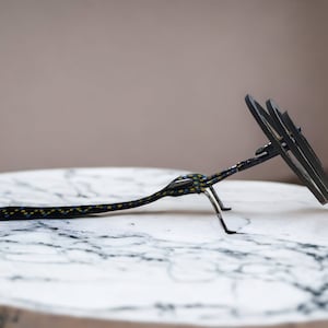 May include: A black metal sculpture with a long, thin body and a circular head. The sculpture is decorated with yellow and blue dots and is sitting on a white marble surface.