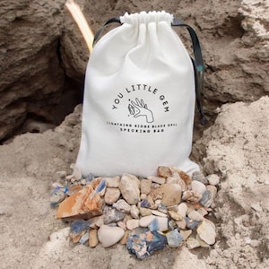 Op de afbeelding: Een witte trekkoordtas met de tekst "You Little Gem Lightning Ridge Black Opal Specking Bag" erop gedrukt. De tas is gevuld met kleine, kleurrijke stenen.