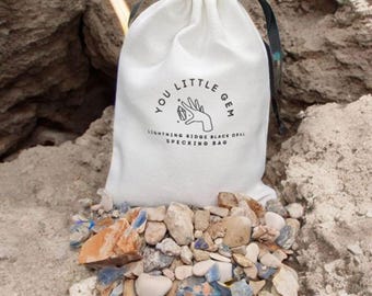Op de afbeelding: Een witte trekkoordtas met de tekst "You Little Gem Lightning Ridge Black Opal Specking Bag" erop gedrukt. De tas is gevuld met kleine, kleurrijke stenen.