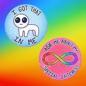 Puede incluir: Dos chapas circulares con acabado brillante. La chapa superior es azul claro con una ilustración de una criatura blanca y el texto I GOT THAT IN ME. La chapa inferior es rosa con un símbolo de infinito arcoíris y el texto ASK ME ABOUT MY SPECIAL INTEREST.