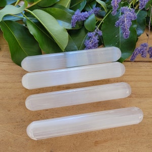 Op de afbeelding: Vier witte selenietstaven, ook wel selenietstokken genoemd, zijn in een rij op een houten oppervlak gerangschikt. De staven zijn glad en gepolijst, met een licht doorschijnende uitstraling.