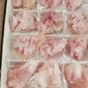 Puede incluir: Una caja de especímenes minerales de color rosa y melocotón. Los cristales tienen una estructura geométrica y en capas y están dispuestos en compartimentos individuales. Los minerales varían en tonalidad, desde rosa pálido hasta un color rosa más intenso. Los cristales se exhiben sobre una envoltura de plástico transparente.