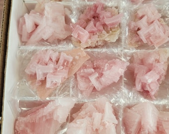 Op de afbeelding: Een doos met roze en perzikkleurige mineraalmonsters. De kristallen hebben een gelaagde, geometrische structuur en zijn gerangschikt in afzonderlijke compartimenten. De mineralen variëren in tint, van lichtroze tot een diepere roze kleur. De kristallen worden gepresenteerd op een transparante plasticfolie.