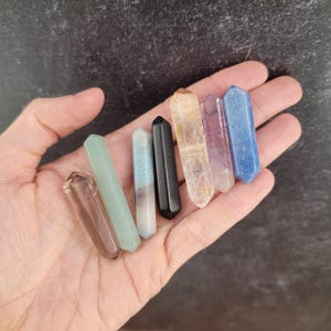 May include: A collection of seven crystal wands in various colors, including brown, green, blue, and black. Each wand is a slender, elongated shape with a pointed tip. The crystals are held in a hand against a dark background.