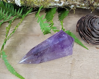 May include: A deep purple amethyst crystal point with a smooth, polished surface. The crystal is shaped like a long, thin pyramid with a pointed tip.