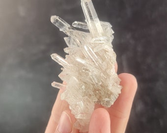 May include: A cluster of clear quartz crystals with a rough, natural texture. The crystals are arranged in a pointed, spiky formation.