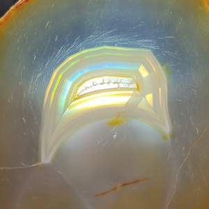 May include: A close-up of a polished agate slice with a rainbow iridescent pattern. The agate has a light blue background with a yellow and white band in the center.